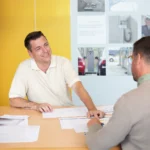 Zwei Männer sitzen an einem Tisch und besprechen ein Angebot einholen gewerblich; einer lächelt, während er auf die Unterlagen zeigt.